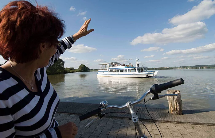 Eine Fahrradfahrerin winkt vom Ufer aus der MS Altmühlsee zu.