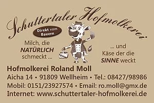 Beige rechteckige Tafel mit brauner Schrift und Bild einer aufrecht stehenden Comic-Kuh in der Mitte.  Im Kopf steht in einem Bogen "Schutterhaler Hofmolkerei". Unter der Kuh ist ein Text, der die Anschrift und Kontaktdaten enthält.