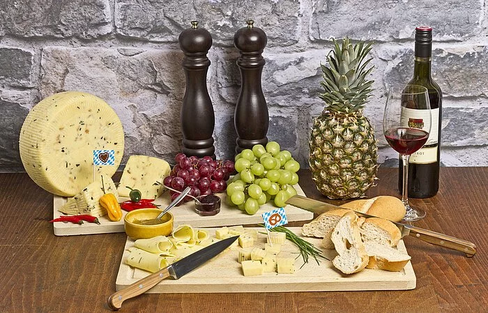 Holzplatte auf der aufgeschnittener Käse liegt, ein Käsemesser im Vordergrund. Hinter der Platte sind von links nach rechts: Flasche Wein, Ananas, zwei große Salz- und Pfeffermühlen in braunem Holz, Weintrauben in weiß und rot und ein runder Käselaib.