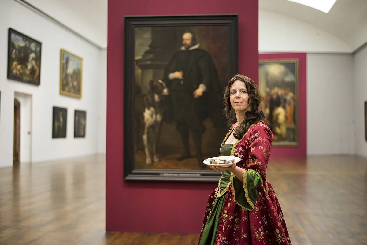 Eine Frau in einem historischem Kleid trägt einen weißen Teller mit Essen in einer Hand. Im Hintergrund ist ein Porträt von Ottheinrich im Residenzschloss zu sehen.