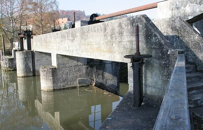 Das massive Betonwehr steht im Mittelpunkt. Rechts sind Treppenstufen hinter einer Mauer zu erkennen. Dahinter kann man vereinzelt ein paar Häuser erkennen.