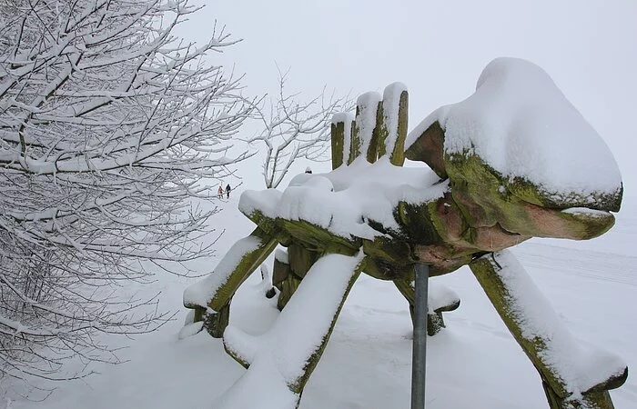 Am Waldrand und im Hintergrund eine Schnee bedeckte Landschaft steht ein Dinosaurier aus Holz neben der Loipe.