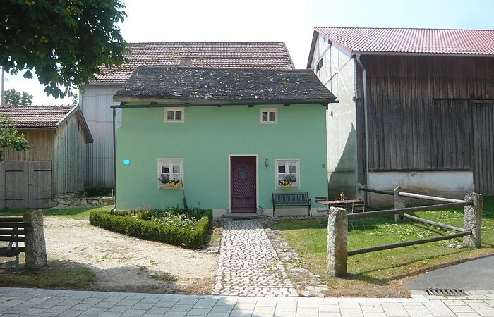 Ein kleines grünes Jurahäuschen mit einem grauen Legschieferdach. Vor dem Häuschen ist ein kleiner Garten daneben jeweils eine Scheune.