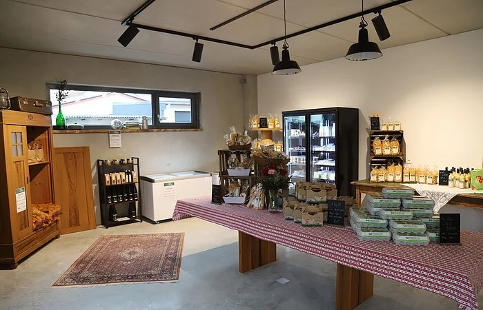 Innenraum eines Selbstbedienungshofladens. In der Mitte ein langer Holztisch mit rotweiß-karierter Tischdecke. Darauf, von links nach rechts, Holzregal mit verschiedenen Nudelpackungen, Vase mit roten u. rosa Nelken, offene Eier in Pappkarton und Eier in Eierschachteln. Im Raum befinden sich noch ein offener Holzschrank mit Kartoffelsäcken, ein Regal mit versch. Produkten, zwei Gefriertruhen, ein Kühlschrank mit Käse-, Wurst- und Milchprodukten. daneben ein kleinerer Tisch mit Nudelpackungen, Eierlikörflaschen und Saftpackungen.