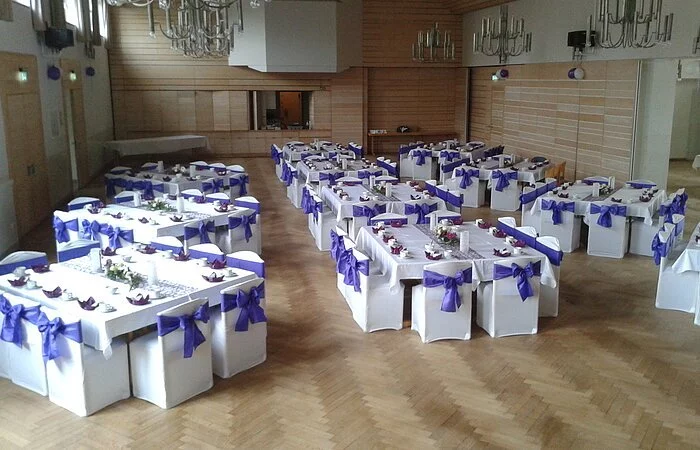 Festliche Bestuhlung in 10er Blöcken, in Lila-weiß, Stühle mit weißen Hussen und dunkel lila Schleifen. Bild von oben geschossen, zeigt den ganzen großen Saal.
