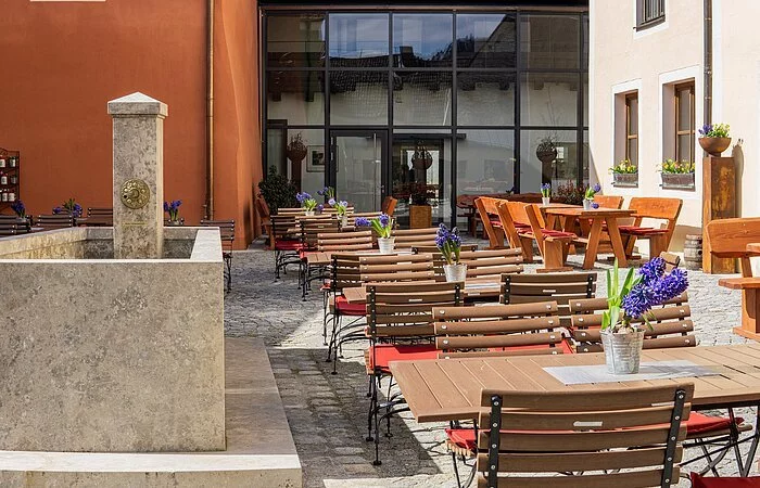 Der Biergarten der Gastwirtschaft Schattenhofer  ist umgeben von den Wänden und dem Neubau im Hintergrund aus Glas. Links ist ein Steinbrunnen zu sehen.
