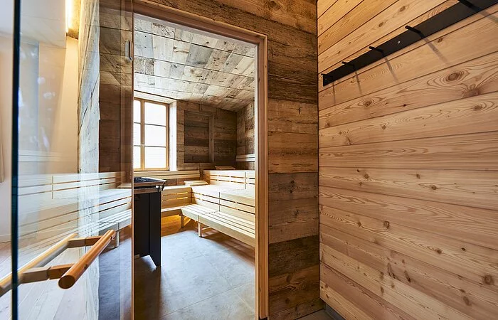 Die Glastür der Sauna aus Holz steht offen und wir können einen Blick auf die leeren Bänke darin werfen. Ein Fenster gegenüber lässt Licht herein.