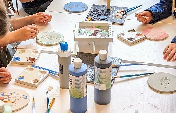 Im Keramikatelier sind auf einem Tisch viele Utensilien, um Keramikrohlinge zu verschönern. Z.B. Farben, Pinsel, Kratzwerkzeuge. Viele Hände sind zudem zu sehen, die gerade ihr Werk verschönern.