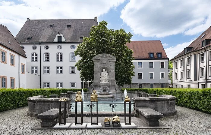Zentrum des Bildes ist eine Ädikula mit einer Steinstatue, die die heilige Maria, die Patrona Bavaria, mit Kind darstellt. Unmittelbar davor befindet sich das rechteckige Brunnenbecken, welches von einer kniehohen Stein-Mauer begrenzt wird.
Dahinter stehen ein großer Baum und niedrige Hecken, die den Kiesplatz, auf dem der Brunnen steht, einsäumen.
Im Hintergrund befinden sich verschiedene mehrstöckige Häuser in hellen Farben.
