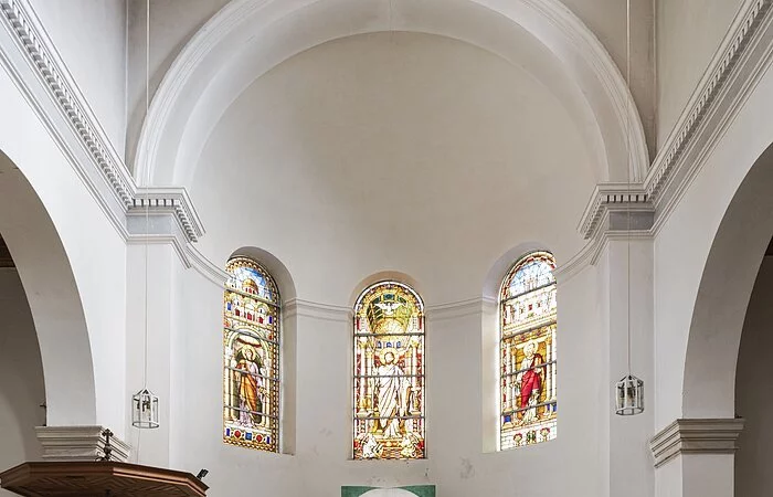 Sie stehen in den Bankreihen der Kirche vor dem Altarraum. Er ist halbrund geformt und die Wände sind weiß. Mittig steht ein großes, modern gestaltetes Kreuz mit Jesusfigur und ein schlichter Altartisch. Darüber sind drei Fenster aus buntem Glas zu sehen, die Petrus, Jesus und Paulus zeigen. Links im Bild befindet sich an einer Säule eine weiße Kanzel mit hölzernem Dach.
Erlöserkirche (Außenansicht)
Das Bild zeigt eine Kirche aus roten Ziegeln. In der Mitte führt eine helle Steintreppe zum Kircheneingang. Über dem Portal befindet sich ein Abbild von Jesus. Auf der linken Seite der Kirche erhebt sich ein Glockenturm mit schwarzem Dach, der fast doppelt so hoch wie die Kirche ist. Darauf steht ein Kreuz.