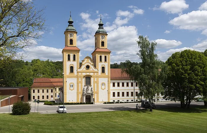 Das Bild zeigt die Klosterkirche St. Johannes der Täufer in Frontalansicht. Zwei barock gehaltene Kirchtürme fassen links und rechts das Mittelschiff und damit auch den Eingang der Kirche. Die Türme sind mehrgeschossig und haben ein Haubendach als Abschluss.  An der rechten Kirchenseite schließt ein langgezogenener Konventbau an. Alle Gebäudeteile sind in ein einem warmen Gelb- und Weißton gehalten. Den Hintergrund des Bildes bildet der langgezogene Bergrücken des Frauenberges.
