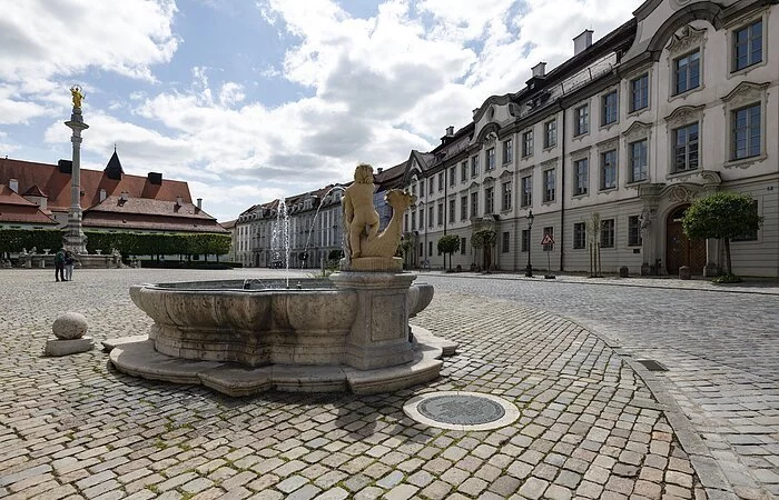 Die Aufnahme zeigt den Residenzplatz, wobei sich der Blick rechts auf eine lange Reihe von mehrstöckigen Häuserfassaden richtet.
Der Residenzplatz selbst ist durchgehend mit Kopfsteinpflaster bedeckt. Im Hintergrund befindet sich ein Brunnen, in dessen Mitte auf einer hohen Säule eine vergoldete Marienstatue thront. Links davon ist die Residenz im Blickfeld, die allerdings nicht vollständig im Bild ist. Unmittelbar vor dem Betrachter steht auf der rechten Bildseite ein zweiter kleiner Brunnen.