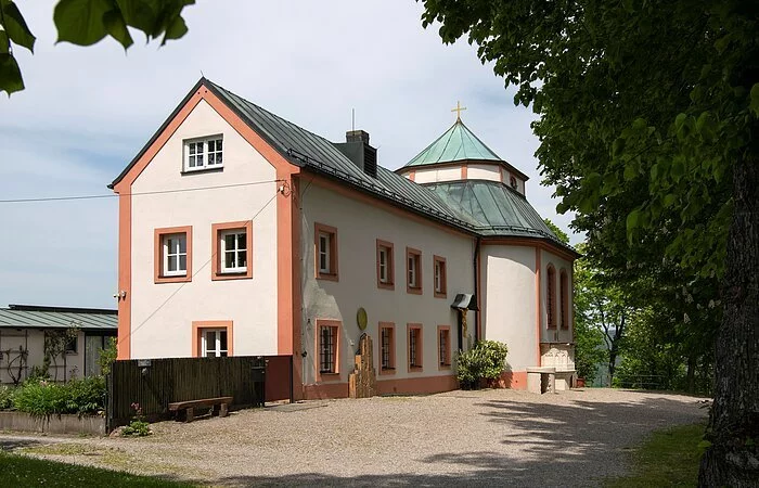 In der Mitte des Bildes steht die Frauenbergkapelle mit angebautem Mesnerhaus. Das Mesnerhaus mit vielen Fenstern und einem Holzkreuz steht dabei im Vordergrund, die schlichte kleine Kapelle im Hintergrund. An der Kapellenseite sind zwei große Fenster und eine Kanzel aus Stein. Das Kapellendach hat eine geknickte Dachfläche mit steilerem Unterdach und flacherem Oberdach. An der Spitze ist ein schlichtes goldenes Kreuz angebracht. Sowohl das Haus, als auch die Kapelle sind cremefarben mit lachsfarbenen Hauskanten und Lisenen. Der Vorplatz ist mit grauem Schotter bedeckt.