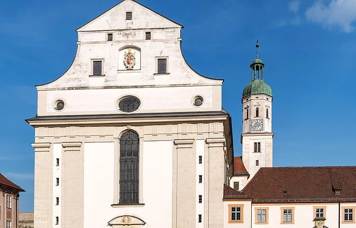 Die Aufnahme zeigt die Fassade der Schutzengelkirche mit einem hohen Fenster über der Eingangstür, rechts daneben liegt das angrenzende Priesterseminar. Direkt vor der Kirche befindet sich der Leonrodplatz. Schräg rechts hinter der Kirche ist ein hoher Turm mit einer Uhr zu erkennen.