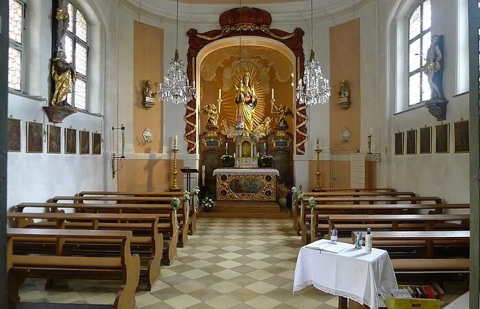 Das Bild zeigt den Altarraum der Frauenbergkapelle. Ein Mittelgang führt auf den Altar zu, auf welchem sich eine hölzerne Madonnenstatue mit dem Jesuskind befindet. Neben der Mutter Gottes sind zwei vergoldete Engel. Die Statuen und der Altar sind von einem rot-goldenen, in Stein gemeißelten Vorhang umrahmt. Die Längsseiten sind jeweils mit zwei Fenstern ausgestattet, zwischen denen sich links ein Kruzifix und rechts eine Statue des Heiligen Sebastian befinden. Unter den beiden Statuen hängen Bilder, die den Kreuzweg zeigen. Links und rechts im Raum stehen Holzbänke.
