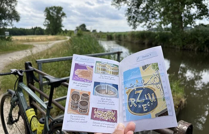 Ein Fahrrad ist an ein Geländer gelehnt. Das Rätselheft der Heimaterlebnistouren ist aufgeschlagen und es sind Abbildungen zu erkennen. Im Hintergrund schlängelt sich ein Radweg durch Wiesen und ein paar vereinzelte Bäume stehen hie und da.