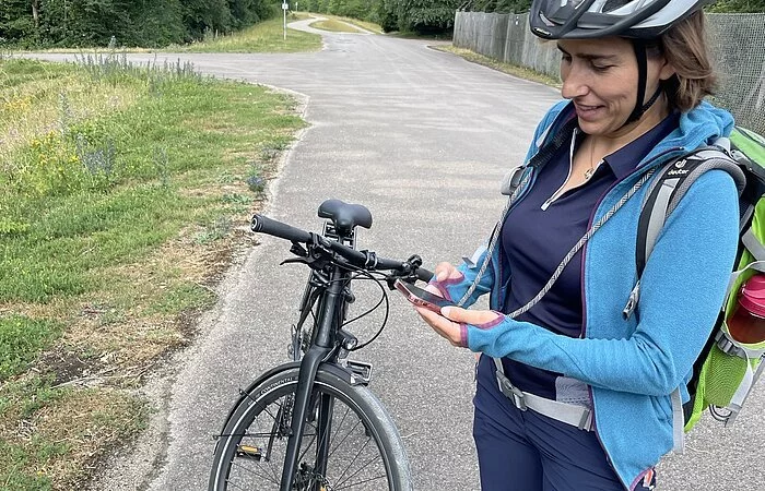 Eine Radfahrerin mit weißem Helm steht neben ihrem Rad auf einem Radweg. Sie hält das Rätselheft in der einen und ihr Smartphone in der anderen Hand, um den nächsten Hinweis zu enträtseln.