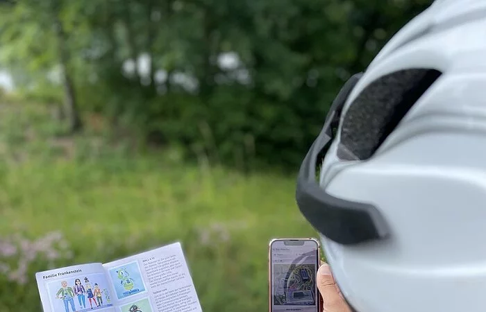 Eine Radfahrerin mit weißem Helm hält das Rätselheft in der einen und ihr Smartphone in der anderen Hand, um den nächsten Hinweis zu enträtseln.
