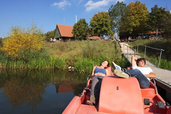 Eine Frau und ein Mann liegen auf einem Tretboot an einem Steg im Altmühlüberleiter. Der steg führt durch eine grüne Böschung hianuf zum Kiosk in Gern.