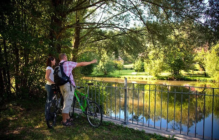 Am Flussufer der Roth in Eisenhammer genießen zwei Fahrradfahrer den einmaligen Ausblick auf die Roth. Die Roth ist an den Ufern umgeben von grünen Sträuchern und Bäumen.