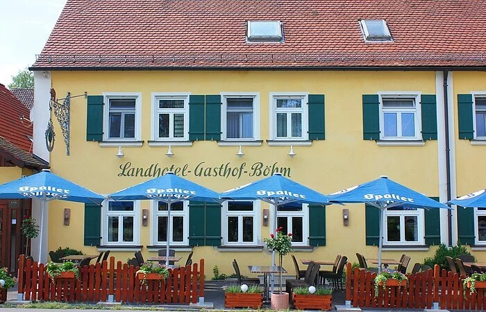 Teilansicht eines zweistöckigen Hauses im fränkischen Baustil mit gelber Fassade und grünen Fensterläden. Davor ein kleiner Biergarten mit braunem Zaun und blauen Sonnenschirmen.