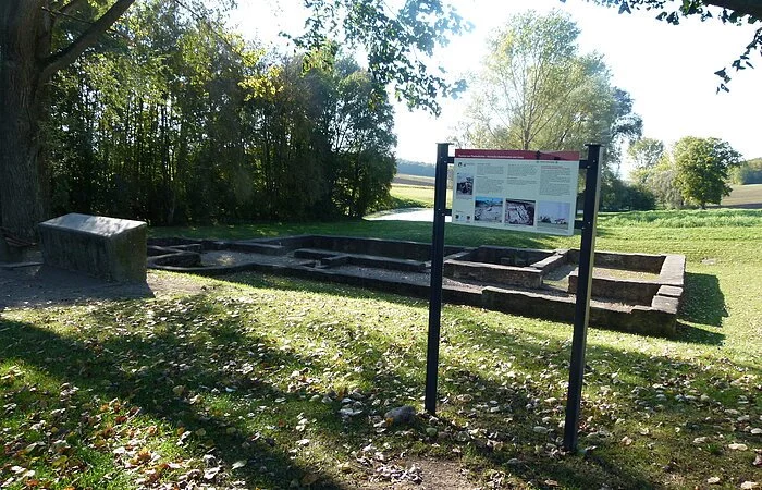Eine römische Ruine auf einer grünen Wiese. Davor steht eine Infotafel.