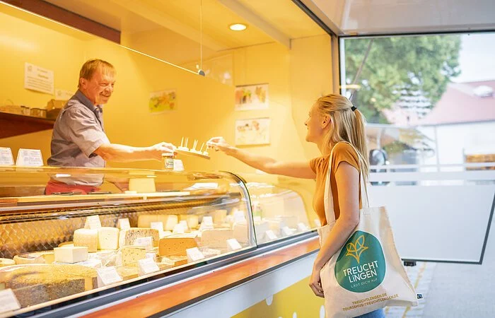 Aufnahme eines Käsestands am Treuchtlinger Wochenmarkt. Eine Frau kauft gerade an der Käsetheke ein. Sie trägt einen Treuchtlinger Stoffbeutel.