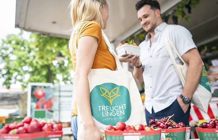 Aufnahme an einem Obst- und Gemüsestand am Treuchtlinger Wochenmarkt. Ein junges Paar steht am Stand neben in Schalen eingepackten Kirschen. Die Frau trägt einen Treuchtlinger Stoffbeutel.
