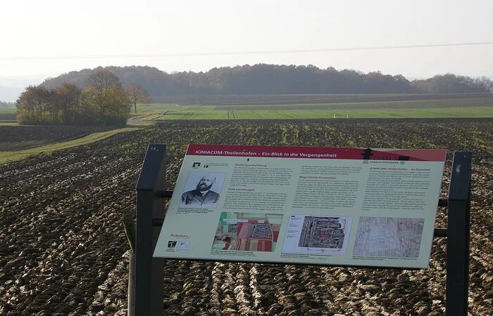 Eine Infotafel steht an einem gepflügten Feld. Im hiintegrund grüne Wiesen und ein Wald.