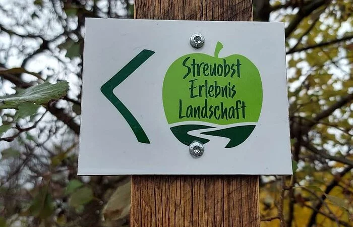 Ein Hinweisschild des Streuobst Erlebnislandschaft, ein grüner Apfel auf weißem Hintergund, hängt an einem Holzpfosten.