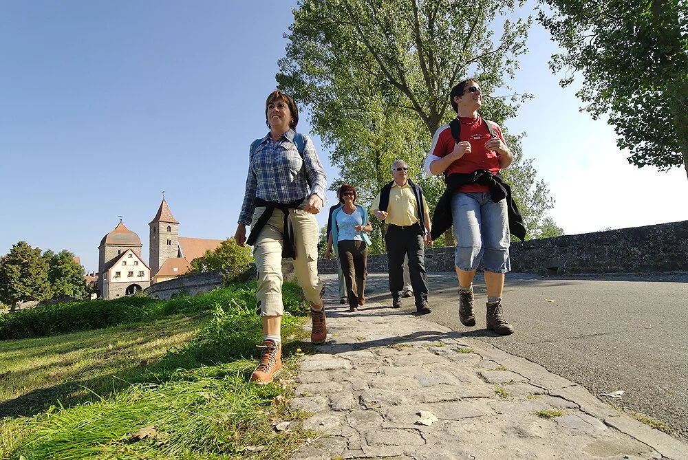 Wanderer wandern über eine Steinbrücke im Grünen. Im Hintergund ein Stadttor.