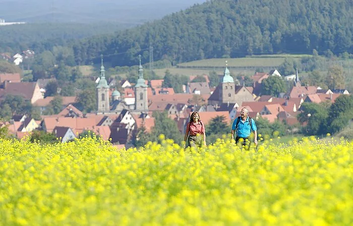 Ein Ehepaar wandert durch ein gelbes Rapsfeld. Im Hintergund die Häuser eines Dorfes.