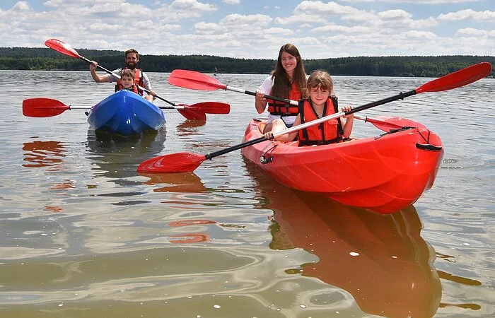 Eine vierköpfige Familie sitzt in zwei Kanus und paddelt über den See. Im blauen linken Kanu sitzt ein Mann mit einem jungen. Im rechten roten Kanu sitzt eine Frau mit einem Mädchen.