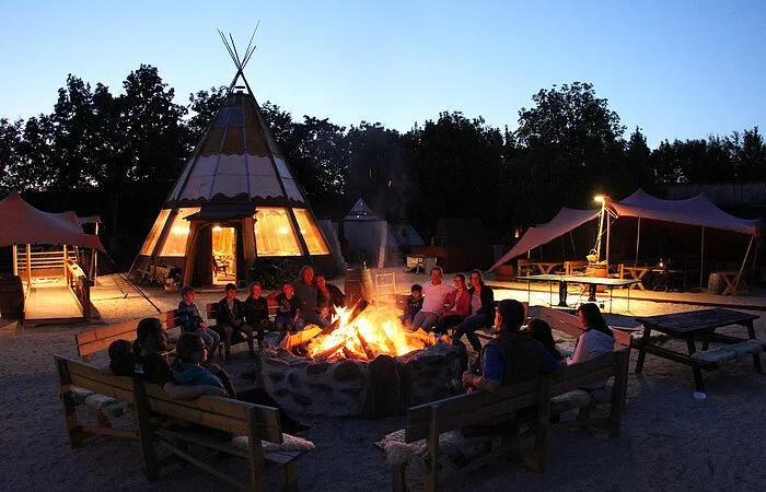 Ein Gruppe Menschen sitzt in der Abenddämmerung auf Holzbänken um ein großes Lagerfeuer. Im Hintergrund stehen ein Tippi und ein Sonnensegel.