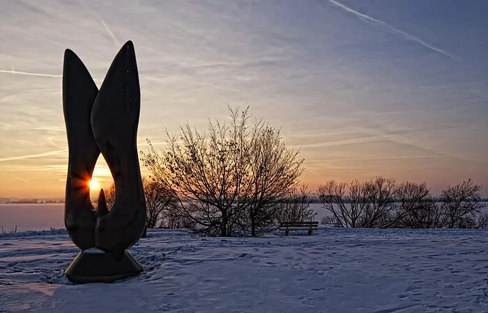 Kunstobjekt bei Sonnenaufgang am schneebedecktem Altmühlsee. Einige Meter dahinter stehen einige Sträucher.