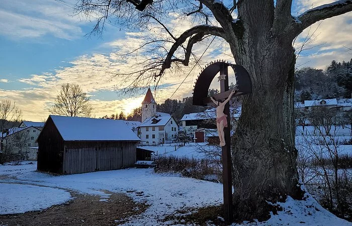 Winterweg mit Baum und einem Kreuz. Im Hintergrund Häuser und eine Kirche mit Schnee bedeckt.