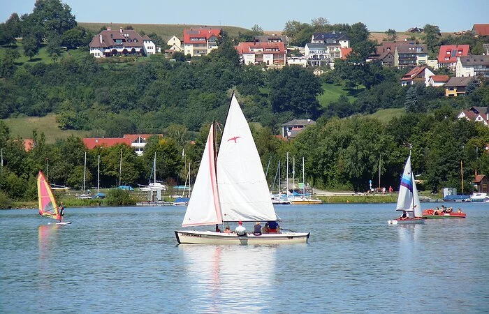 Ein weißes Segelschiff auf einem See. Daneben Windsurfer. Im Hintergund am Ufer Bäume. Etwas weiter hinten Wohnhäuser.