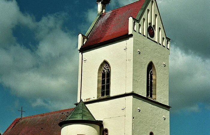 Ein Kirche mit weißer Fassade. Neben dem Kirchenschiff ein großer Kirchturm mit rotem Dach und Storchennest auf dem Schlot.