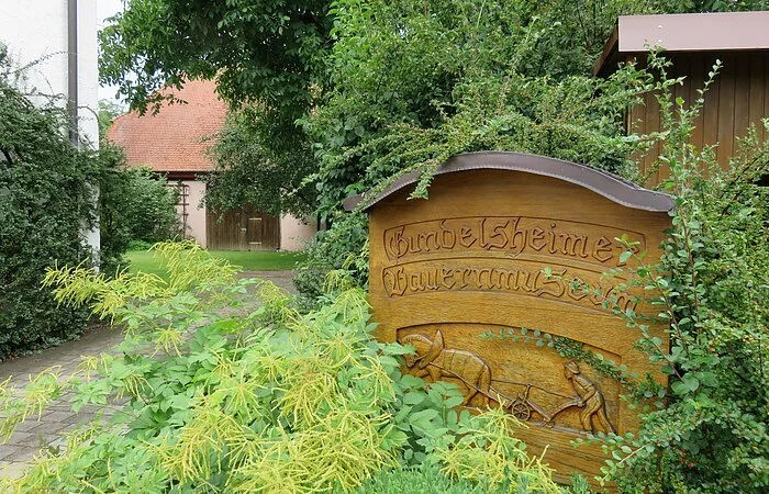 Ein Holzschild umrahmt von grünen Sträuchern und Bäumen. In das Schild sind die Worte Gundelsheimer Bauernmuseum geschnitzt. Links führt eine Straße in einen Hof mit Scheune.