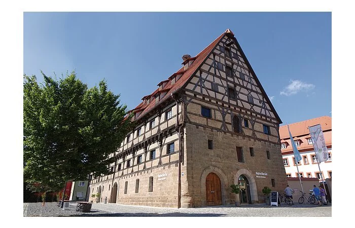 Ein großes Fachwerkhaus mit 6 Ebenen. Die Fassade der unteren zwei Stöcke besteht aus Stein, darüber braunes Fachwerk mit weißer Fassade.