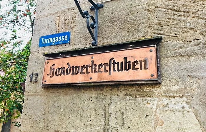 Ein Schild aus Kupfer. Drauf steht Handwerkerstuben. Das Schild hängt an einer Steinmauer. Darüber ein kleines blaues Straßenschild mit der Aufschrift Turmstraße.