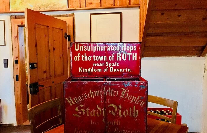 Eine rote Kiste aus Metall steht auf eibnem Tisch in einem Wohnraum. Auf der Kiste steht in weißer Schrift: Unsulphurated Hops of the town of Roth near Spalt Kingdom of Bavaria