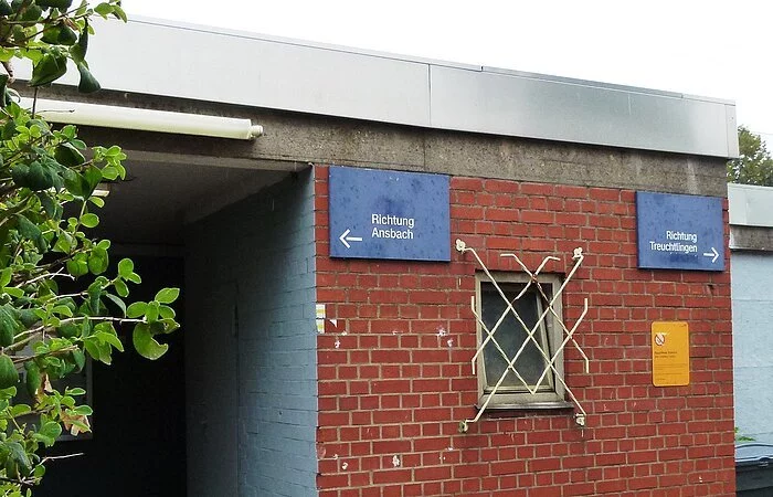 Ein kleines Backsteingebäude mit zwei blauen Schildern und einem Flachdach. Links ein blaues Schild mit den Worten Richtung Ansbach und einem Pfeil nach links. Rechts ein blaues Schild mit den Worten Richtung Treuchtlingen und einem Pfeil nach rechts.