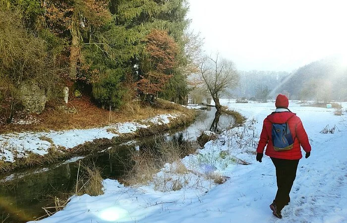 Eine Person wandert auf dem schneebedeckten Weg der Schlaufe 24, neben der fließenden Schambach entlang.