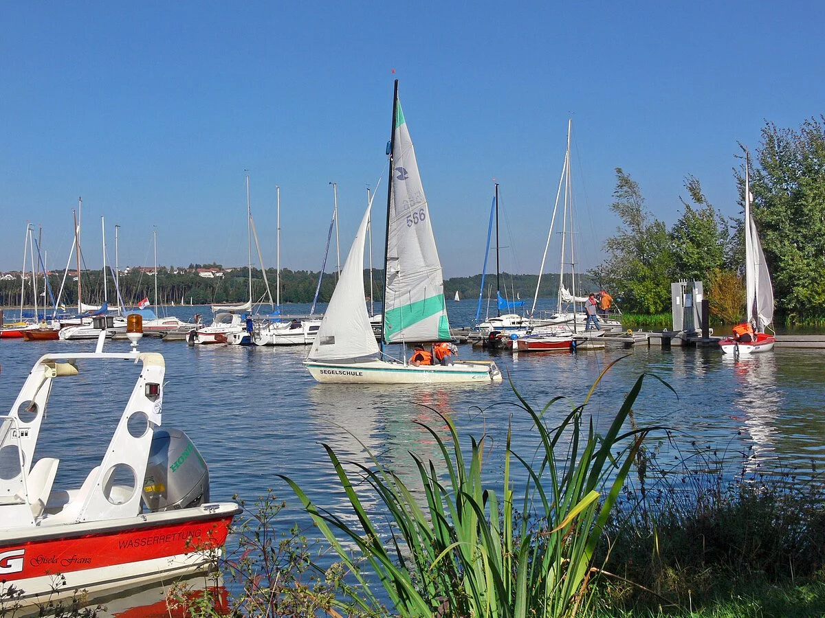 Ein Segelboot segelt gerade aus einem Segelhafen. Im Hafen liegen noch weitere Boote vor Anker.