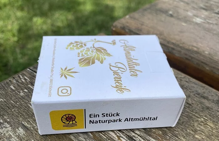 Auf einem vergilbten Holzbrett liegt die Verpackung einer Seife. Weißer Karton, quadratisch, das Logo "Ein Stück Naturpark Altmühltal" ist auf der schmalen Seite angebraucht und zeigt im Bild nach vorne. Auf der flachen Vorderseite ist in goldener Schrift "Altmühltaler Bierseife" eingedruckt, darunter goldene Hopfenblüten. Im Hintergrund sieht man noch einen grünen Rasen.