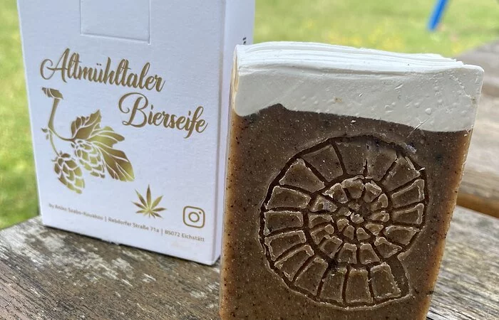 Auf einem Holzbrett steht hochkant die Verpackung, davor die Bierseife, rechteckig, braun mit eingestanztem Ammoniten. Der obere Teil der Seife ist weiß und wellig, wie ein Bierschaum. Auf der weißen Verpackung steht in goldener Schrift "Altmueltaler Bierseife" mit Hopfenblütenabbildung. In Hintergrund sieht man unscharf einen Garten mit Rasen.