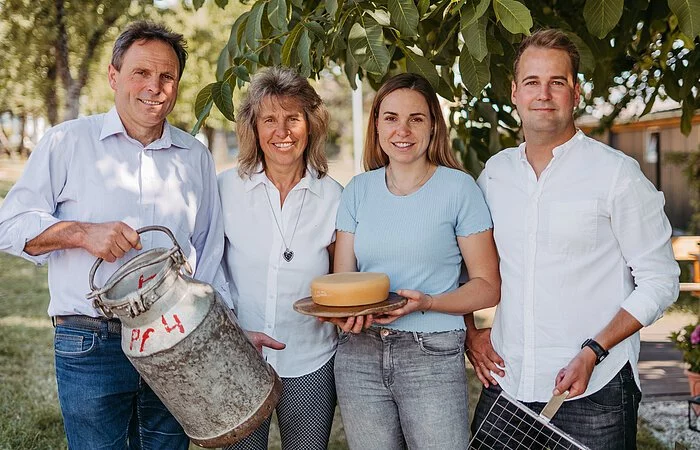 Vier Personen stehen nebeneinander. Mann, Frau, Frau, Mann. Der Mann ganz links hält eine große Blechmilchkanne in der Hand, die zweite Dame ein Holzbrett mit einem Käselaib und der rechte Mann ein Eisengitter zum Käseschöpfen. Alle stehen unter einem Walnussbaum. von dem m Hintergrund die Blätter sichtbar sind.