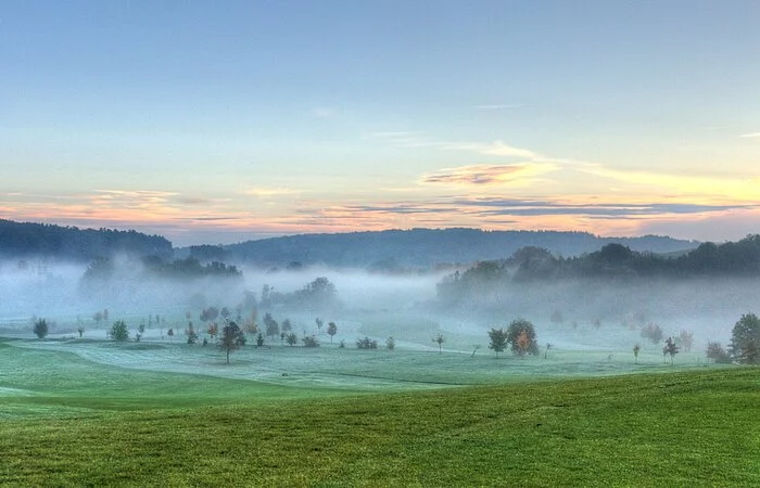 Eine Panorama-Aufnahme einer grünen weiten Wiese. Im Hintergrund Berge. Über die Wiese steht ein Nebelschleier. Der Horizont ist orange verfärbt.