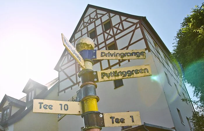 Blick von unten auf einen Schildermast mit goldenen Schildern. Darauf steht in der Reihenfolge von oben nach unten: Drvingrange, Kurzplatz, Puttingarden Tee 10 und Tee 1. Im Hintergund ein großes Fachwerkhaus mit braunen Balken